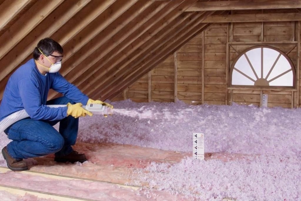 A man in a blue shirt and jeans uses a spray gun to apply pink insulation in a construction setting.