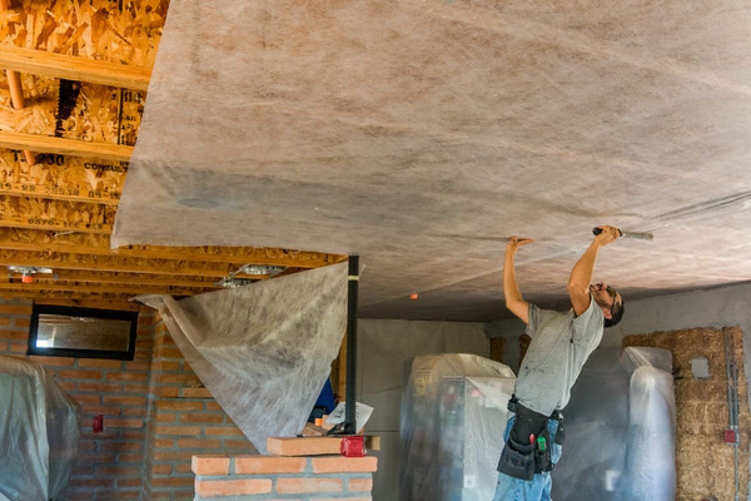 A man is working on the ceiling of a house, focused on his task while standing on a ladder with tools in hand.