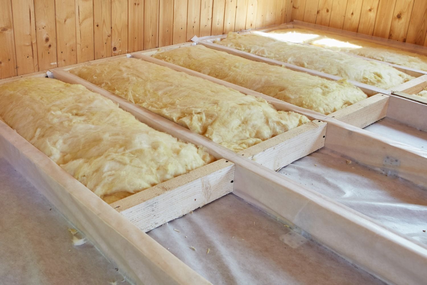 Insulation batts installed in wooden frames on a floor, with a wooden wall in the background.