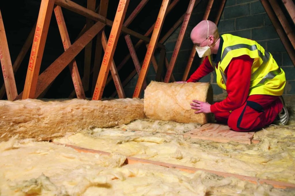 A man wearing a yellow vest and safety gear installs insulation in an attic, focusing on his task in a well-lit space.
