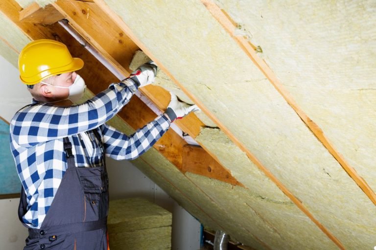 A man wearing a hard hat and overalls installs insulation in an attic, focusing on his work in a well-lit space.
