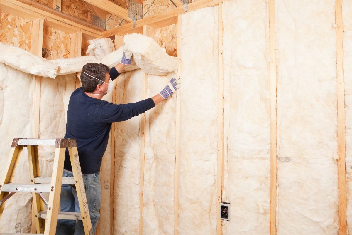 A person installs insulation batts in a wooden wall frame while standing on a ladder, surrounded by construction materials.