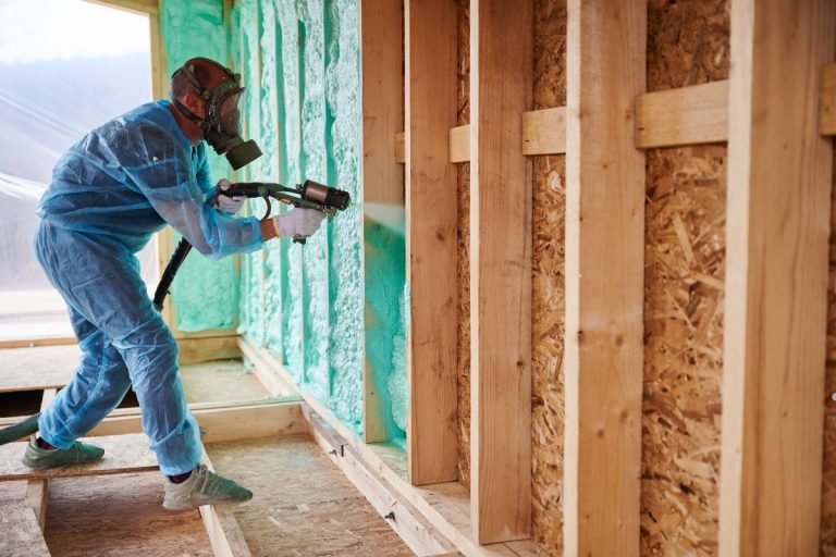 A worker in protective gear applies spray foam insulation to the wooden frame of a house under construction.